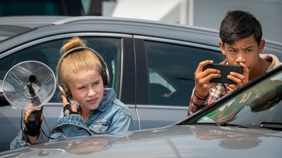 Pippa und Jet - versteckt hinter einem Auto - testen die neue Lippen-Ablese-App von Jet © NDR/Letterbox Foto: Boris Laewen