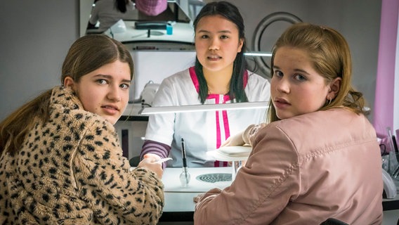 Lou und Pippa sitzen an einem Tisch im Nagelstudio, auf der anderen Seite des Tisches sitzt Mai. © NDR/Letterbox Foto: Boris Laewen