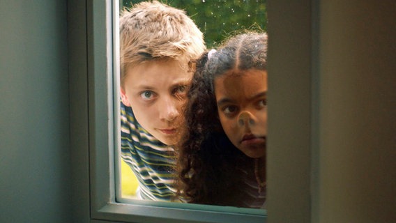 Moritz (Jan Albatross) und Amy (Melody Rose) schauen von außen durch eine Fensterscheibe und drücken sich an der Fensterscheibe die Nasen platt. © NDR/Letterbox 