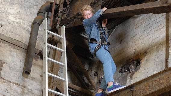 Max (Bruno Alexander) steht mit einem Klettergurt auf einem Stahlträger. © NDR/Studio Hamburg Foto: Romano Ruhnau