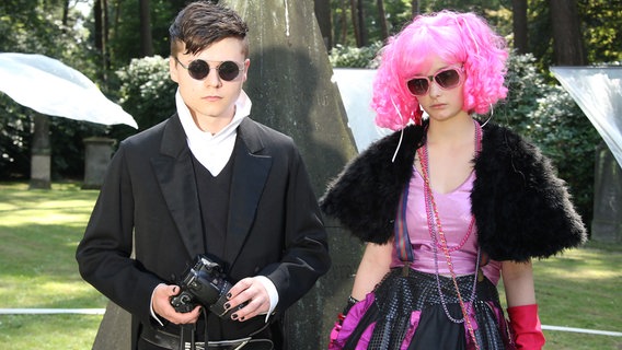 Ein Junge (Tom Böttcher) und ein Mädchen (Johanna Werner) mit Sonnenbrillen und auffälliger Kleidung stehen unter Bäumen. © NDR/Studio Hamburg Foto: Romano Ruhnau