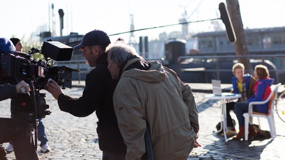 Die Kamera ist auf Max und Tatja gerichtet, die an einem Tisch am Hafen sitzen. © NDR Foto: Claudia Timmann