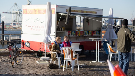 Max und Tatja sitzen an einem Tisch am Hafen. Die Tonangel ist auf sie gerichtet. © NDR Foto: Claudia Timmann