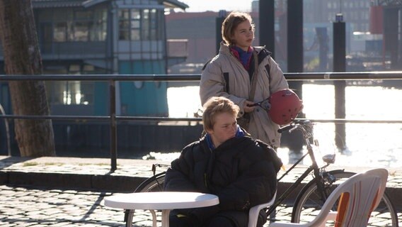 Max sitz an einem Tisch am Hafen. Tatja steht hinter ihm und hält einen Helm in der Hand. © NDR Foto: Claudia Timmann