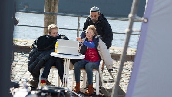Max und Tatja sitzen am Hafen an einem Tisch. Sie haben sich warme Jacken umgelegt. Ein Mann mit Mütze steht hinter Tatja. © NDR Foto: Claudia Timmann