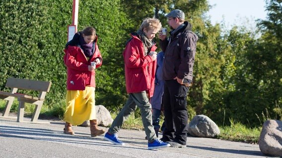 Nina und Max gehen in warmen Jacken mit einem Becher in der Hand auf einer Straße. Zwei Menschen stehen im Hintergrund. © NDR Foto: Claudia Timmann