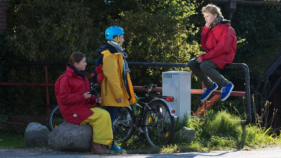 Nina und Jessi schauen zu Max, der vor ihnen auf einem Geländer sitzt. Alle drei tragen dicke Jacken. © NDR Foto: Claudia Timmann
