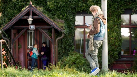 Max lehnt mit seinem Handy in der Hand an einem Laternenpfahl. Im Hintergrund stehen Jessi und Nina an einer Haustür. © NDR Foto: Claudia Timmann