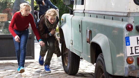 Jessi und Max schleichen geduckt hinter einem einem Geländewagen © NDR Foto: Claudia Timmann