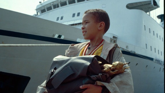 Ein Junge steht vor einem großen Schiff am Kai und hat goldene Statuen in einer Tasche © NDR 