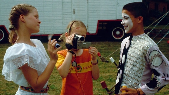 Natascha und Vivi interviewen Rafael mit einer Videokamera vor einem Zirkuswagen © NDR 