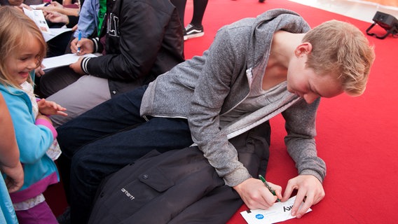 Bruno schreibt sitzend sein Autogramm auf einen Zettel. Ein kleines Mädchen steht vor ihm. © NDR Foto: Claudia Timmann
