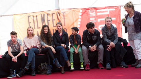 Pfefferkörner sitzen nebeneinander auf der kleinen Bühne im Filmfestzelt, die Kinderbetreuerin steht daneben. © NDR Foto: Claudia Timmann