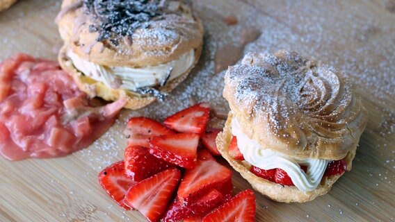 Zwei Windbeutel gefüllt mit Vanille-Creme und Schokoladensahne. Daneben liegen Rhabarberkompott und geschnittene Erdbeeren. © NDR Foto: Florian Kruck