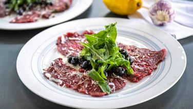 Carpaccio mit Heidelbeeren, Parmesan und Rucola auf einem Teller angerichtet. © NDR Foto: Frank von Wieding