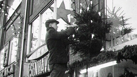 Ein Mann befestigt einen Weihnachtsbaum an einem Schaufenster. Ein Mann befestigt einen Weihnachtsbaum an einem Schaufenster. © NDR Foto: NDR