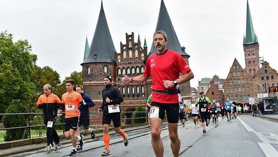 Ein Läufer joggt mit hochgestrecktem Daumen vor dem Holstentor in Lübeck entlang, in Hintergrund sind weitere Läufer zu sehen. © PRO EVENT Foto: Roman Klawun