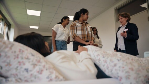 Eine Trainerin erklärt einer Gruppe Frauen an einem Krankenbett Arbeitsschritte. © NDR 