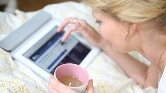 Eine junge Frau liegt auf einem Bett und bedient mit der einen Hand ein Tablet und hält in der anderen hand einen Becher Kaffee. © NDR Foto: Berit Ladewig