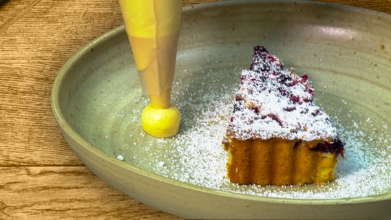 Ein Stück Brombeerkuchen mit einem Klecks Verbene-Creme auf einem Teller angerichtet. © NDR 
