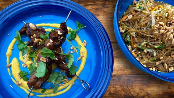 Entenbrust-Spieße mit Mango-Currysoße und Glasnudelsalat auf blauen Tellern angerichtet. © NDR Screenshot/Montage 