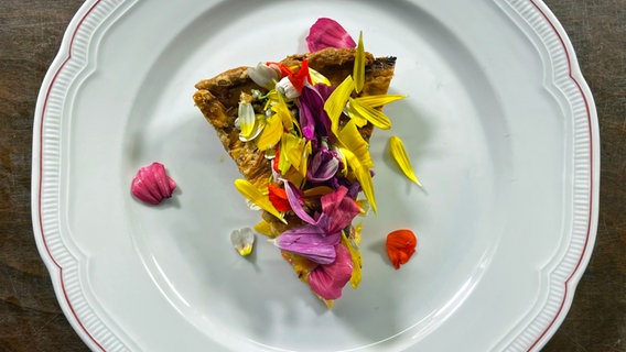 Ein Stück Tomatentarte mit essbaren Blüten dekoroert auf einem Teller serviert. © NDR 