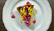 Ein Stück Tomatentarte mit essbaren Blüten dekoroert auf einem Teller serviert. © NDR 
