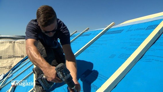 Ein Dachdecker schraubt auf einem Dach eine Planke fest. © NDR 