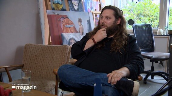 Künstler Justus von Karger sitzt bei einem Interview auf einem Sofa in einem Zimmer. © NDR 