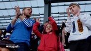 Ein Mann (l.), ein Mädchen (m.) und ein Junge (r.) stehen auf einer Tribünes eines Fußballstadions auf den Färöer Inseln und klatschen in die Hände. © NDR 