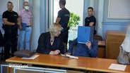 Ein Angeklagter sitzt beim Prozessauftakt im Fall Drogenhandel in Norderstedt in einem Gerichtssaal und hält eine blaue Mappe vor seinem Gesicht. © NDR 