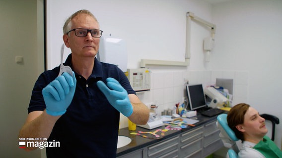 Ein Zahnarzt steht in seinem Arztzimmer mit einem kleinen Bumerang in der Hand. © NDR 