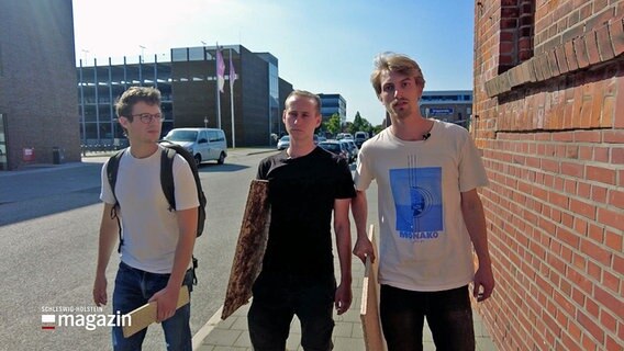 Drei junge Männer gehen an einer Straße lang und tragen dabei Bauplatten. © NDR 