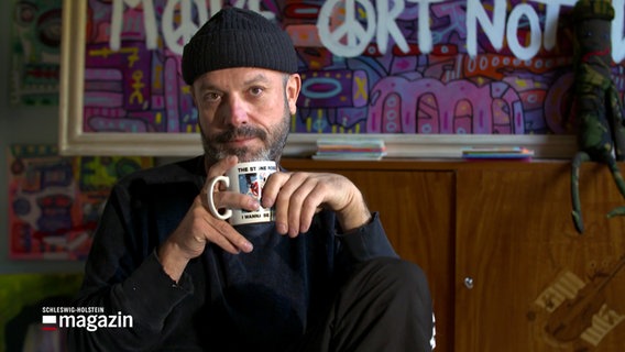 Künstler Jan Plewka sitzt in einem Raum mit einer Tasse in den Händen und blickt nachdenklich in die Kamera. © NDR 