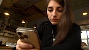 Eine junge Frau sitzt an einem Tisch in einem Cafe und blickt auf ihr Smartphone. © NDR 