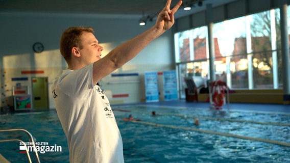 Schwimmtrainer Alexander Müller steht in eine Schwimmhalle in Lübeck und zeigt mit zwei Fingern die Anzahl der noch zu schwimmenden Bahnen an. © NDR 