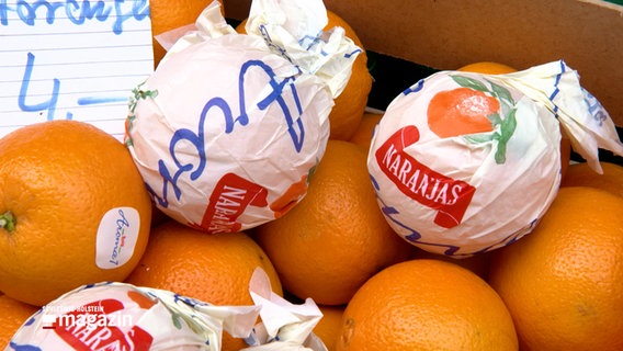 Zwei in Papier gewickelte Orangen liegen auf anderen losen Orangen auf einem Tisch auf einem Wochenmarkt in Kiel. © NDR 
