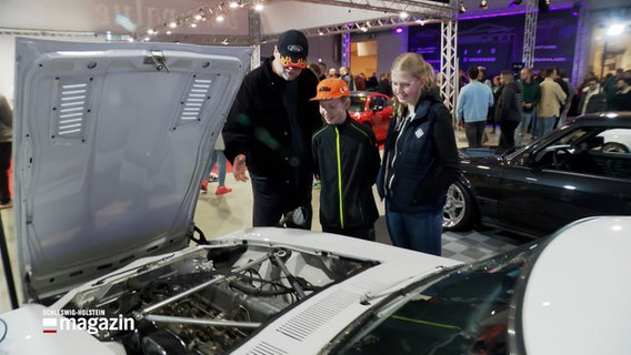 Ein Mann (l.) zeigt mit seiner Hand auf den Motor eines Autos in der Halle der  Nordic Motor Show Messe in Neumüsnter, ein Junge (m.) und ein Mädchen (r.) schauen dabei zu. © NDR 