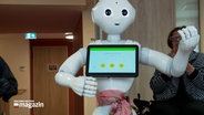 Ein weißer Roboter mit einem Display vor der Brust steht mitten in einem Gruppenraum des Lübecker Pflegezentrums Travetal. © NDR 