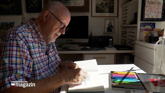 Künstler Manfred Schröder sitzt an einem Schreibtisch und malt etwas in einem Heft. © NDR 