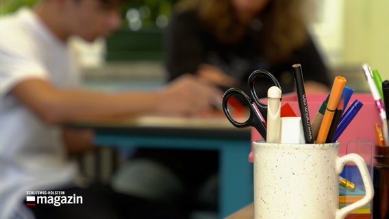 Eine Tasse mit Stiften und einer Schere steht in einem Klassenzimmer. © NDR 