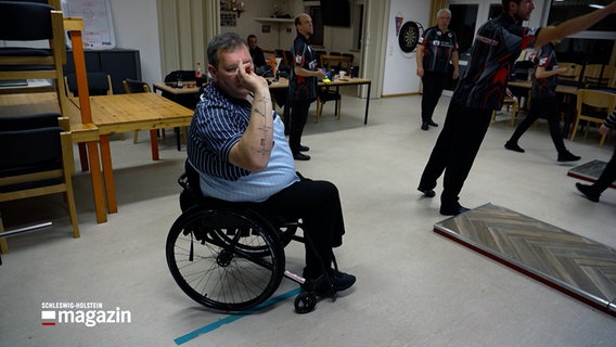 Ein Rollstuhlfahrer spielt Darts in einem Raum mit anderen Spielern. © NDR 