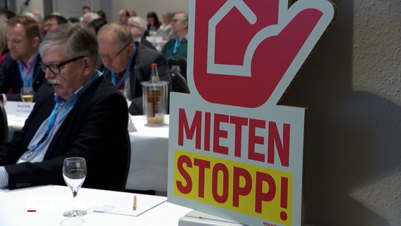 Ein Schild in Form eine Handfläche mit dem darunterstehenden Schriftzug Mieten Stopp! steht auf einem Tisch in einem Konferenzraum. © NDR 