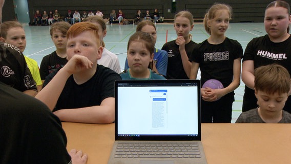 Ein Laptop steht vor einer Gruppe Kinder in einer Sporthalle. © NDR 