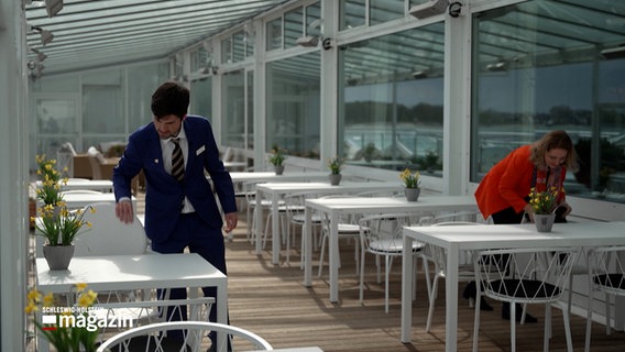 Ein junger Mann in einem Anzug richtet einen Tisch auf der Terrasse eines Restaurants ein. © NDR 