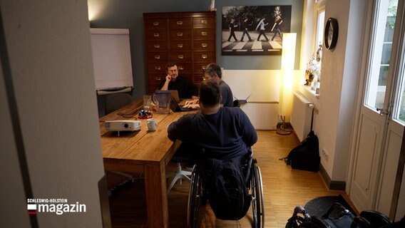 Ein Rollstuhlfahrer sitzt in einem Büro an einem Tisch mit zwei anderen Menschen. © NDR 