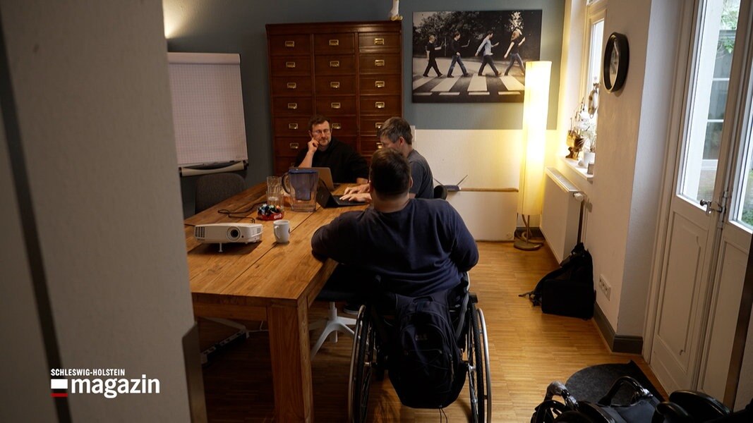 Ein Rollstuhlfahrer sitzt in einem Büro an einem Tisch mit zwei anderen Menschen.