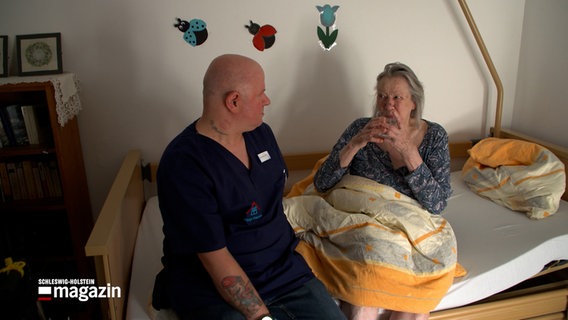 Ein Pfleger sitzt am Bettrand einer Demenz-Erkrankten, die aus einem Becher trinkt. © NDR 