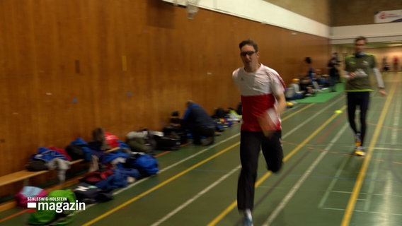 Ein Mann rennt mit einem Staffelstab durch eine Sporthalle. © NDR Foto: NDR Screenshots