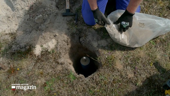 In einem Loch steckt eine tief versenke Metallstange. Dabei handelt es sich um einen Höhenmesspunkt. © NDR Foto: NDR Screenshots